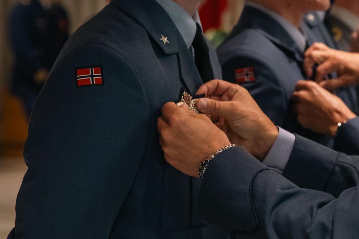 Lähikuva norjalaissotilaista rivissä. Kuvassa näkyy käsiä, jotka kiinnittävät norjalaisten univormun rintapielukseen norjalaista sotilasilmailumerkkiä. Kuvasta on rajattu pois ihmisten kasvot.