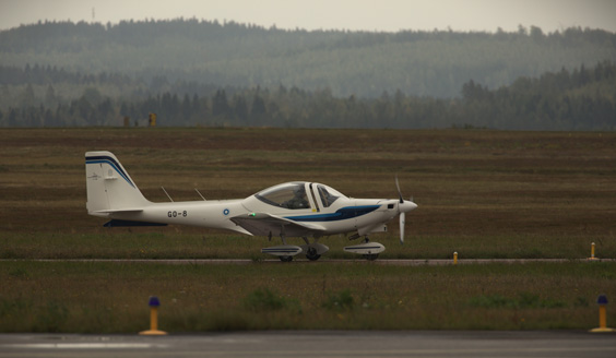 Grob G115E -alkeiskoulukone yksin lentokentällä rullaamassa, taustalla metsäinen maisema.