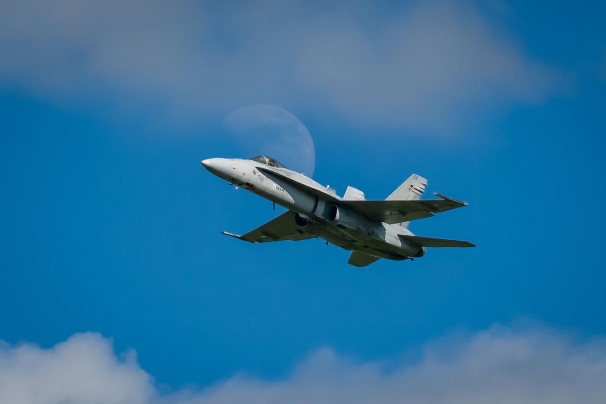 Ilmavoimien F/A-18 Hornet -hävittäjä kirkkaan sinisellä taivaalla. Koneen ylä- ja alapuolella on hentoa pilveä ja sen takana häilyy kuu,