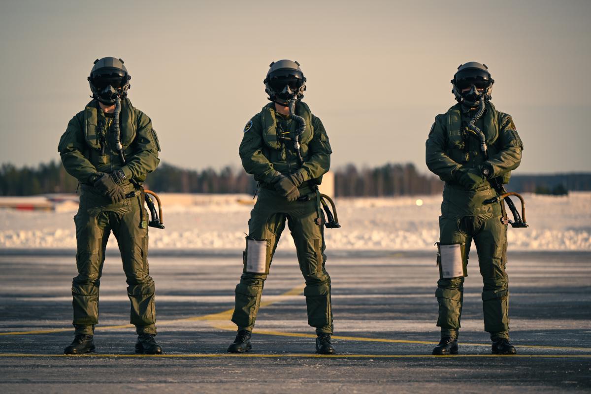 Kolme lentovarusteisiin pukeutunutta henkilöä seisoo rivissä rintamasuunta kameraan päin. Kuva on otettu lentokentän alueella talvella, ja lentokypärien visiirit ovat alhaalla.