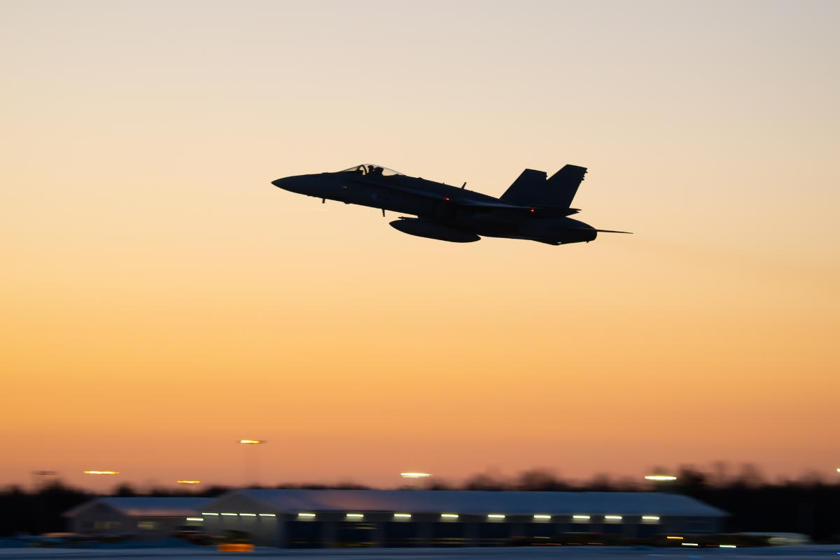 F/A-18 Hornet -monitoimihävittäjä lentää oranssiksi värjäytyneellä taivaalla. Koneesta näkyy vain musta siluetti.