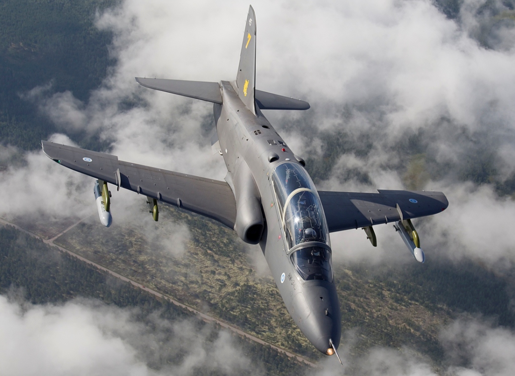 Kuvassa ilmavoimien harmaa Hawk Mk 51 -suihkuharjoituskone lentää pilvisellä taivaalla. Koneen siipiripustimiin on kiinnitetty savupodit. Pilviverhon välistä näkyy hieman maata.