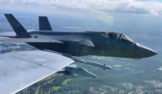 Suomalaisen Hornet-hävittäjän ohjaamosta otettu kuva yhdysvaltalaisesta F-35-hävittäjästä, joka lentää Hornet-hävittäjän siivellä kesäisessä maisemassa. F-35-hävittäjän alapuolella lentää toinen suomalainen Hornet-hävittäjä.