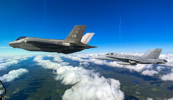 Hävittäjän ohjaamosta otettu kuva, jossa näkyy vasemmalla yhdysvaltalainen F-35-hävittäjä ja oikealla suomalainen Hornet-hävittäjä lentämässä kesäisellä kelillä rakoilevan pilvipeitteen yllä.