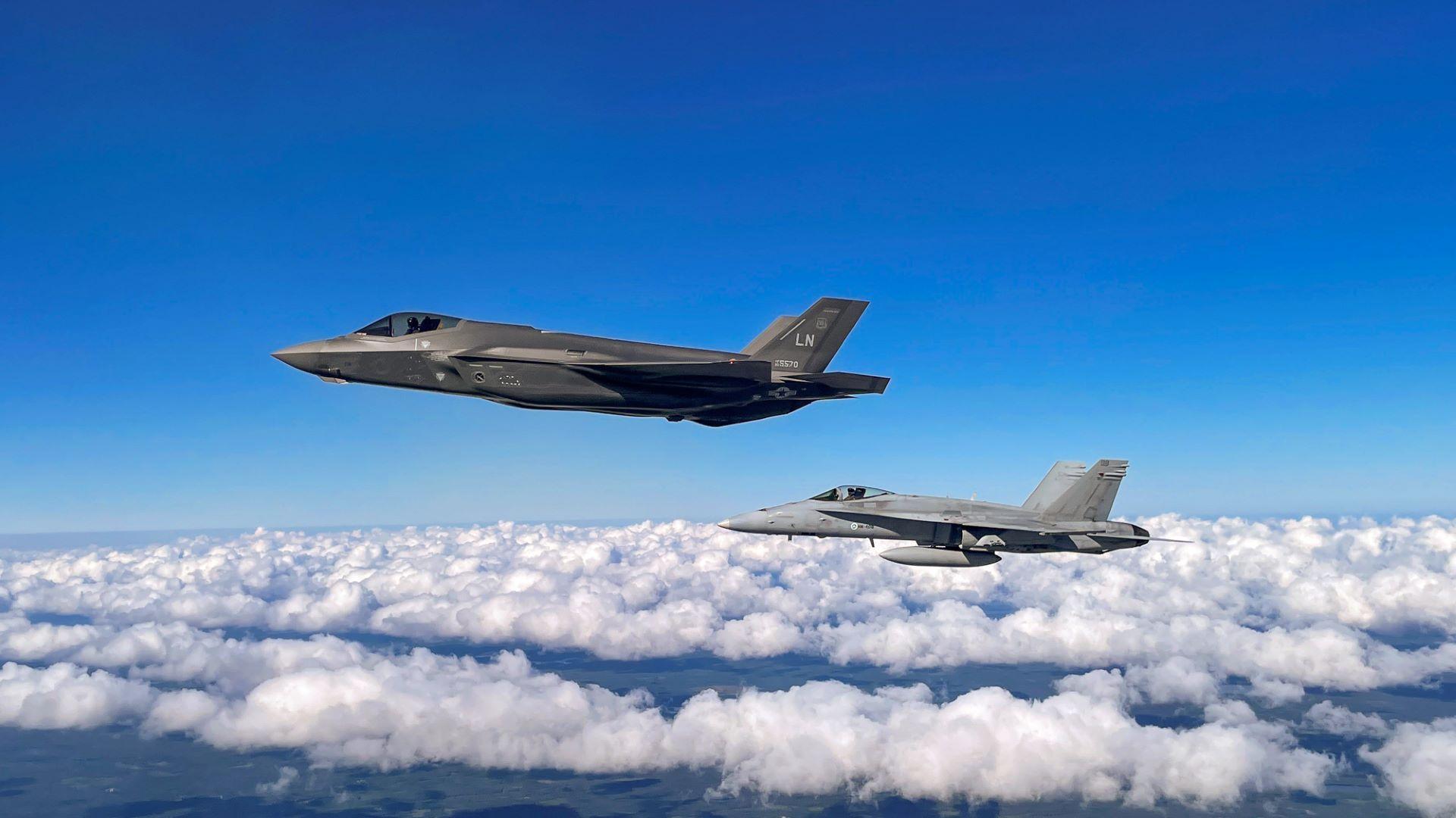 Ilmassa otettu kuva, jossa vasemmalla yhdysvaltalainen F-35-hävittäjä ja oikealla hieman alempana suomalainen Hornet-hävittäjä lentämässä kesäisellä kelillä hieman rakoilevan pilvipeitteen yllä.