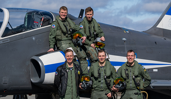 Midnight Hawksin neljä lentäjää ja yksi selostaja poseeraavat harmaan, Suomen lipun värein kuvioidun, Hawk-suihkuharjoituskoneen edessä aurinkoisena päivänä.. 