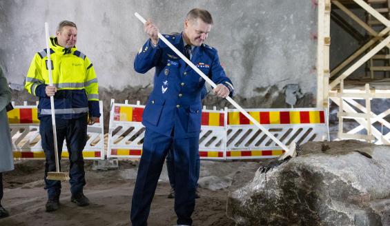 Ilmavoimien komentaja tervaa pitkävartisella harjalla suurta kiveä, ja Puolustuskiinteistöjen toimitusjohtaja seisoo vieressä harjaa pidellen odottaen omaa vuoroaan.