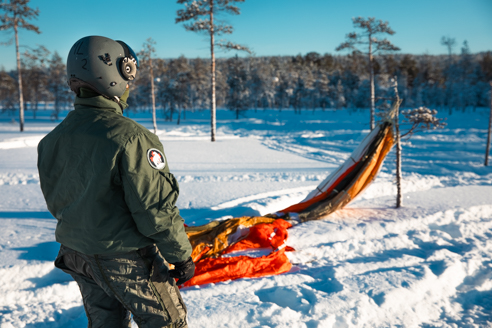 Lentäjä lumisessa maisemassa selin katsojaan, edessään kokoon painunut laskuvarjo.