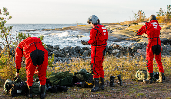 Kolme ohjaajaoppilasta punaisessa suoja-asussa syksyisen meren rannalla.