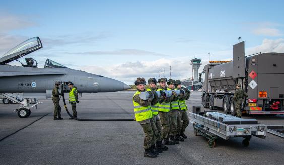 Viisi reserviläistä seisoo rivissä, sylissään yksi ohjus. Reserviläiset valmistautuvat kantamaan ohjuksen yhdessä taustalla näkyvään Hornet-hävittäjään aseistaakseen koneen. 