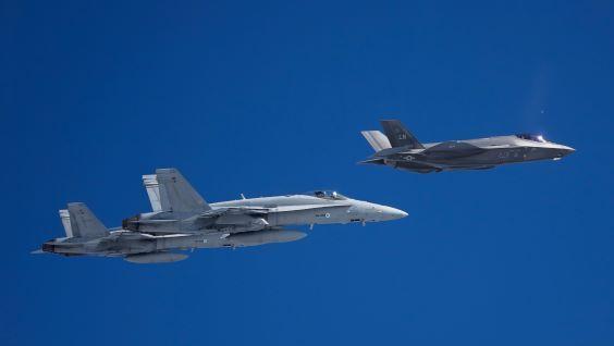 Lentokoneiden sivupuolelta otettu kuva, jossa yhdysvaltalainen F-35 lentää kahden suomalaisen Hornetin edellä sinisellä taivaalla. 