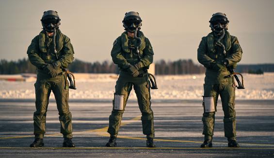 Kolme norjalaista lento-oppilasta seisoo ryhdikkäästi rivissä. He ovat pukeutuneet lentovarusteisiinsa aina kypäriä myöten, kypärien visiirit alhaalla. Lentäjät seisovat lentokenttäalueella, taustallaan kevättalvinen luminen maisema. 