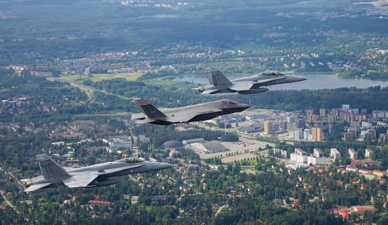 Ilmassa, viistosti hävittäjien yläpuolelta, otetussa kuvassa kaksi Hornetia, välissään F-35, lentää kesäisen Tampereen yllä. Tampereesta näkyy asutusta, puustoa, järvi sekä horisonttiin ulottuvaa metsää.