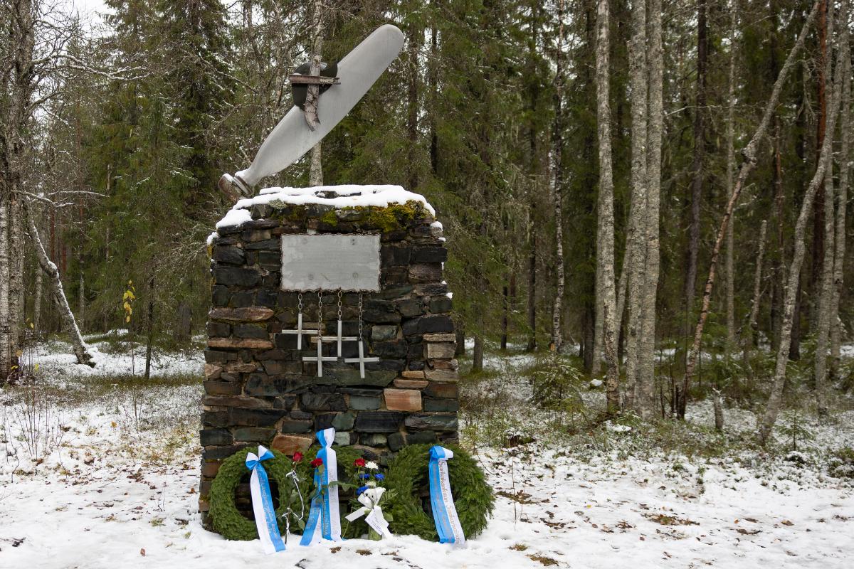 Kivestä ja metallista tehty neliskanttinen muistomerkki lumisessa metsässä. Muistomerkin edessä on havuseppeleitä.