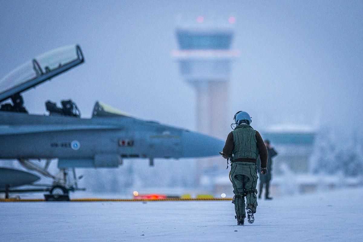 Lentäjä kävelee lumisella platalla kohti taustalla häämöttävää Hornet-hävittäjää ja lennonjohtotornia.