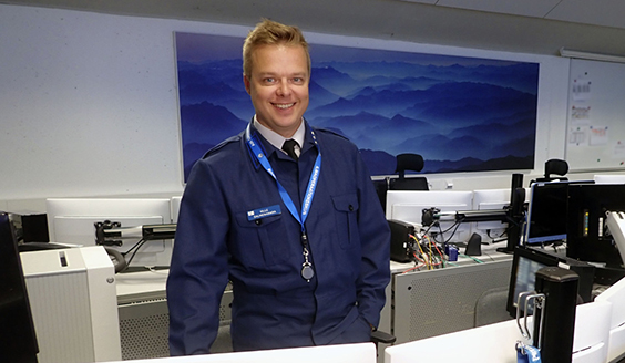 Kapteeni Ville Salmenhaara seisomassa hymyillen työpöydän ääressä. Taustalla on tietokoneen näyttöjä ja taulu.