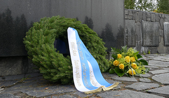 Seppele nojaamassa Lentäjien patsaaseen. Seppeeleessä on sinivalkoiset nauhat, joissa on tekstiä. Vieressä on kimppu keltaisia ruusuja. Taustalla näkyy patsaassa olevia nimiä.