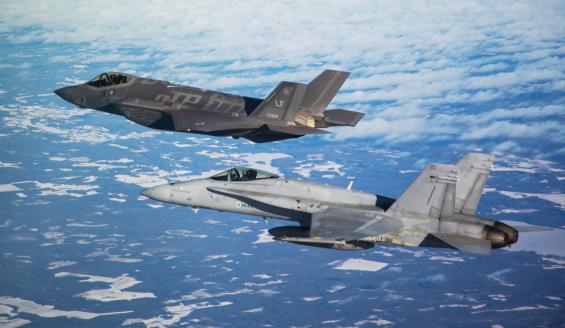 Lockheed Martin F-35A Lightning II ja Suomen nykyinen F/A-18 Hornet -monitoimihävittäjä lentävät rinnakkain ja taustalla sinistä taivasta ja vaaleita pilvenhattaroita